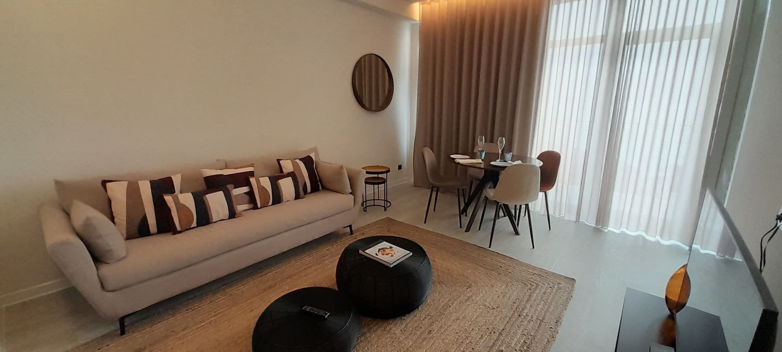 A modern living room with a sofa, ottomans, a dining table with chairs, and a large window with curtains.