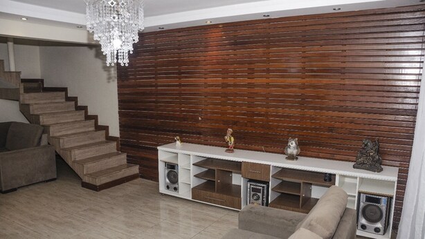 A modern living room with a wooden wall panel, a white entertainment unit, a staircase, and a chandelier. - MANSION SALE NEW (Europa Belo Horizonte)