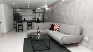 A modern living room with a grey sofa, a glass coffee table, and a kitchen area in the background.