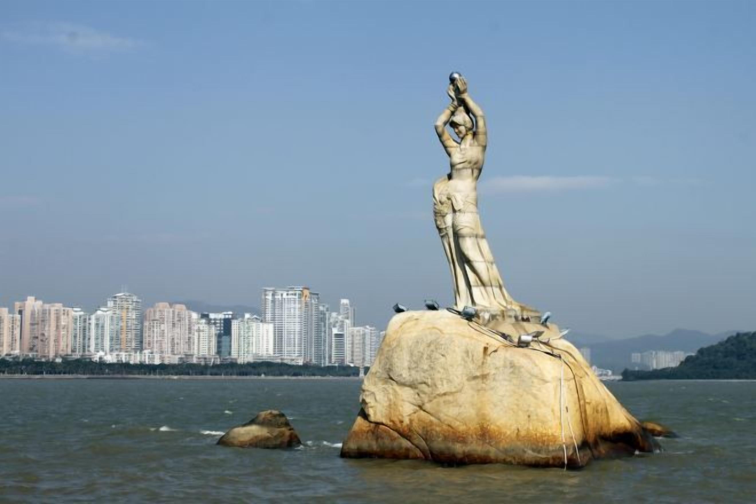 A large rock with a statue on top, surrounded by water and a city skyline in the background.