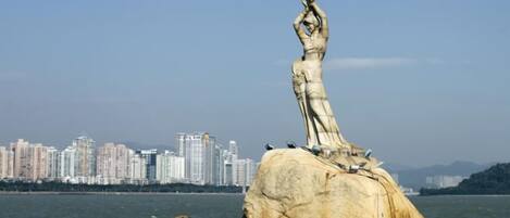 A large rock with a statue on top, surrounded by water and a city skyline in the background.