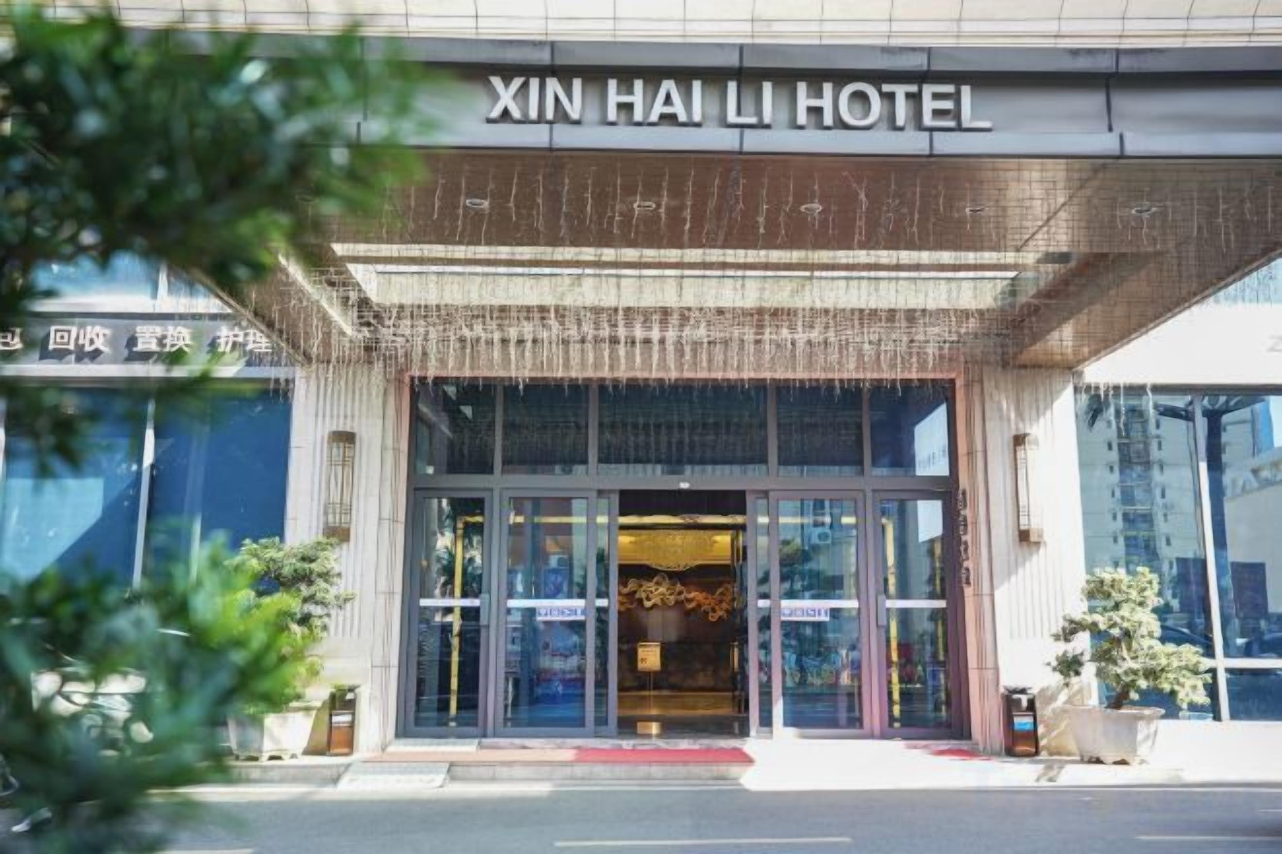 A entrada do Hotel Xin Hai Li, com fachada moderna, grandes portas de vidro e um tapete vermelho que leva à entrada.