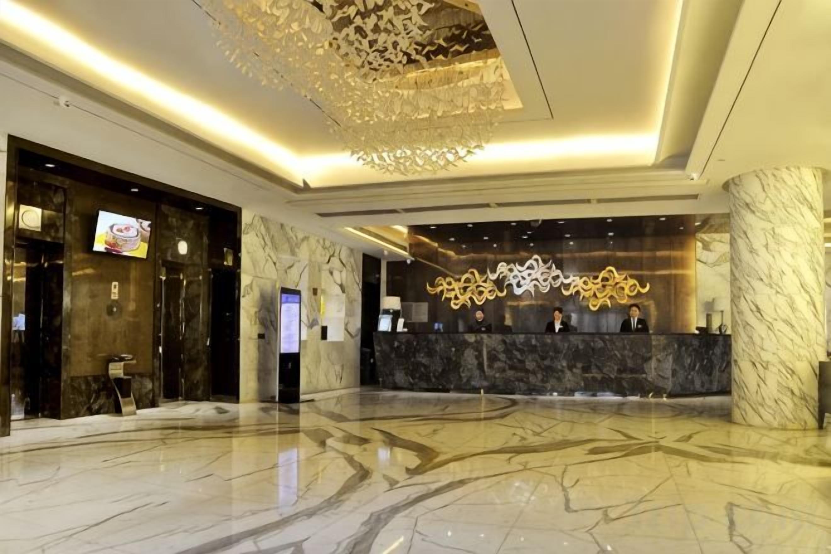 Um hall de hotel com balcão de recepção de mármore, um grande candelabro e uma tela digital.
