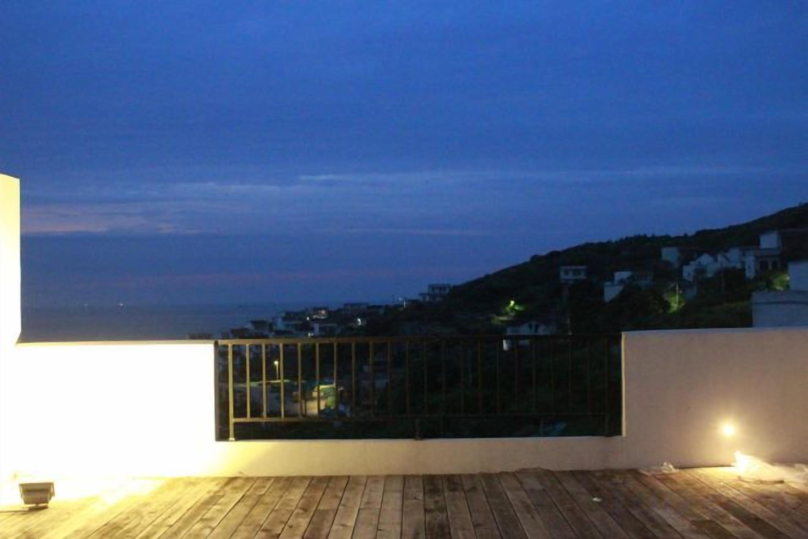 Una terraza en la azotea con suelo de madera, barandilla de cristal y vistas a un pueblo costero al atardecer.