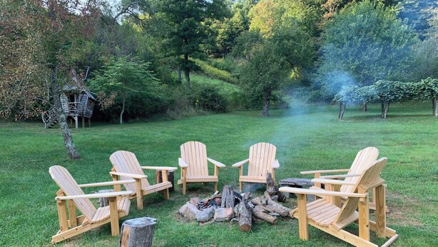 A group of Adirondack chairs arranged around a fire pit in a grassy outdoor area with trees in the background. - Bikers' Loft on the Creeper Trail (Damascus)