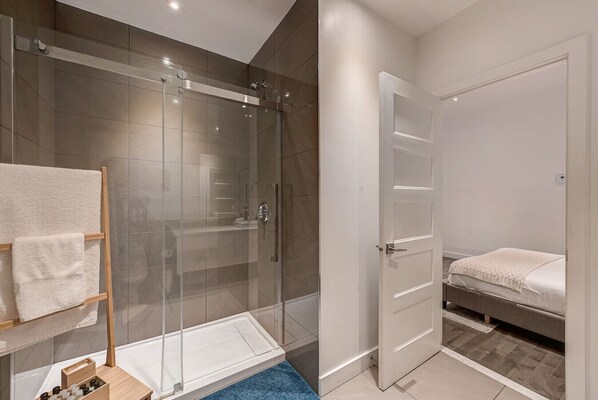 A modern bathroom with a glass-enclosed shower, a white door, and a bed with a blue rug.