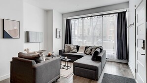 Una sala de estar moderna con un sofá gris, un sillón marrón y una mesa de centro. Hay un ventanal con cortinas, un televisor de pantalla plana montado en la pared y un cuadro enmarcado sobre el sofá.