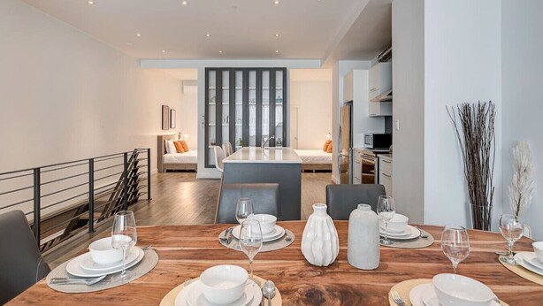 Una cocina moderna con una isla central, una zona de comedor con mesa de madera dispuesta para cuatro personas, y un salón con sofá y televisor.