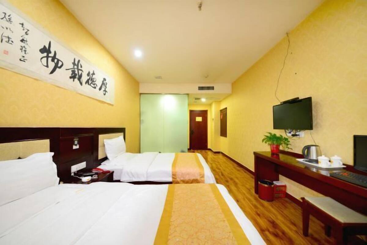 A hotel room with two beds, a desk with a TV, a plant, and a wooden floor.