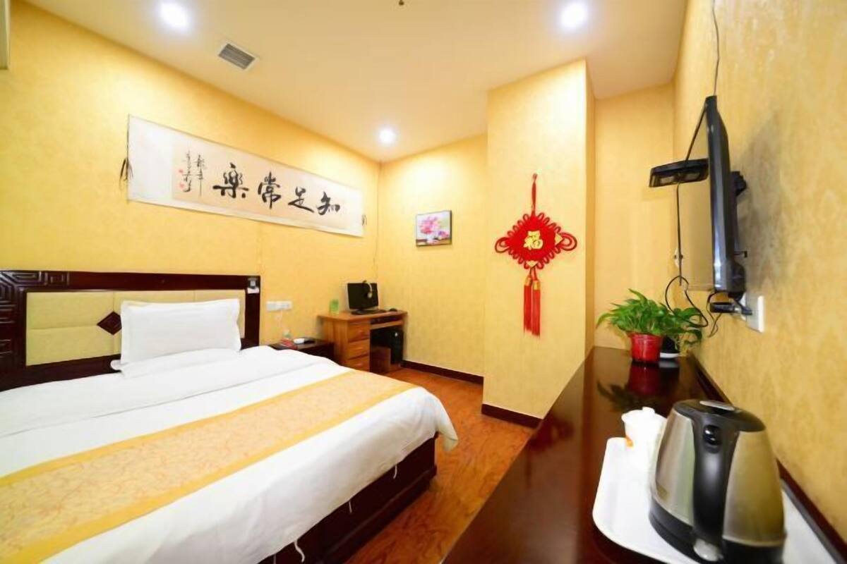 A hotel room with a neatly made bed, a desk with a computer, a TV mounted on the wall, and a red decorative item on the wall.