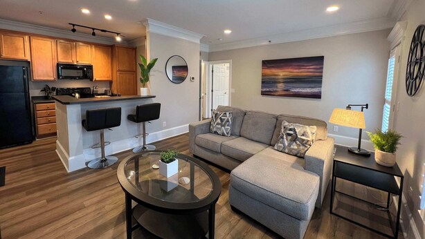 A modern living room with a sofa, ottoman, and coffee table, a kitchen with wooden cabinets and appliances, and a dining area with a bar. - 1 Bdrm|Pet Friendly|North Hills|Long Stays (Raleigh)