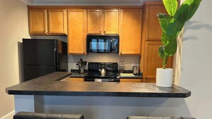 A kitchen with wooden cabinets, a black refrigerator, a microwave, a stove, and a sink. There are two black chairs and a potted plant. - 1 Bdrm|Pet Friendly|North Hills|Long Stays (Raleigh)