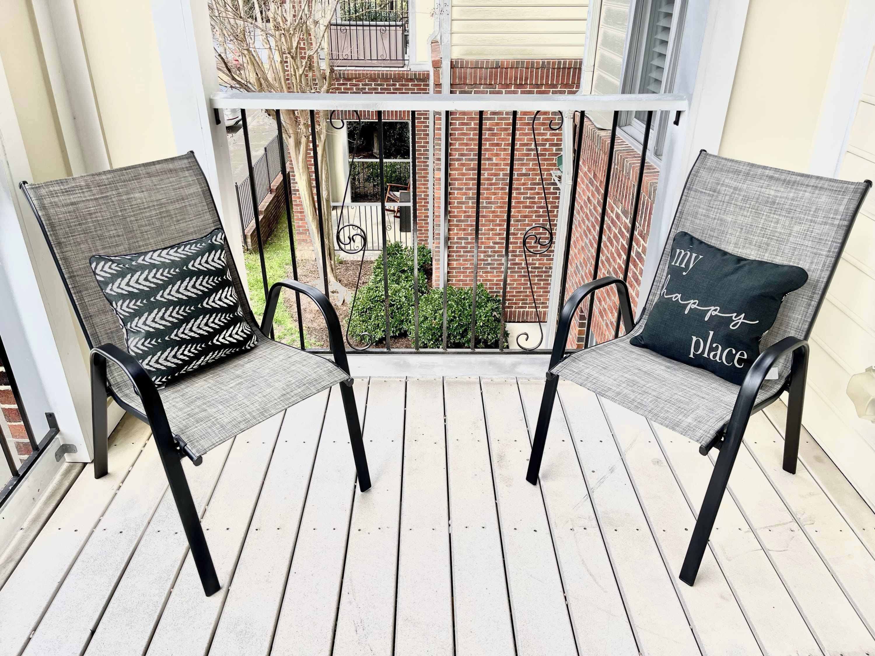 Two outdoor chairs with patterned cushions on a wooden deck, overlooking a brick building with a balcony.