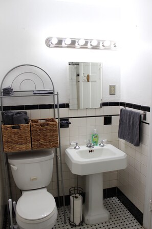 Un baño con inodoro, lavabo y espejo. Hay cestas en un estante sobre el inodoro y una toalla colgada en la pared.