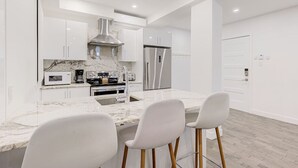 Une cuisine moderne avec des meubles blancs, un plan de travail en marbre, une hotte de cuisinière en acier inoxydable, un réfrigérateur et un four encastré. On trouve deux tabourets de bar blancs au niveau de l’îlot central.