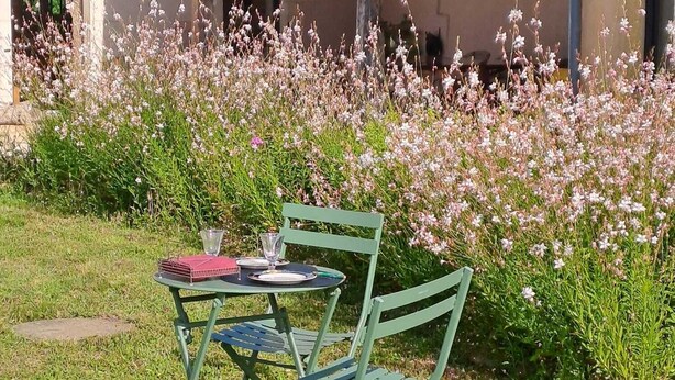 Een groene metalen tafel met stoelset buiten, met een klein tafelkleed en een rood notitieboekje, omringd door een tuin met roze bloemen.