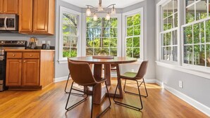 A kitchen with wooden cabinets, a built-in microwave, and a stove. There is a round dining table with two chairs. Large windows provide a view of greenery outside. - Fenced In|Pets OK|By DT Apex|King Bed|EV Charger (Apex)