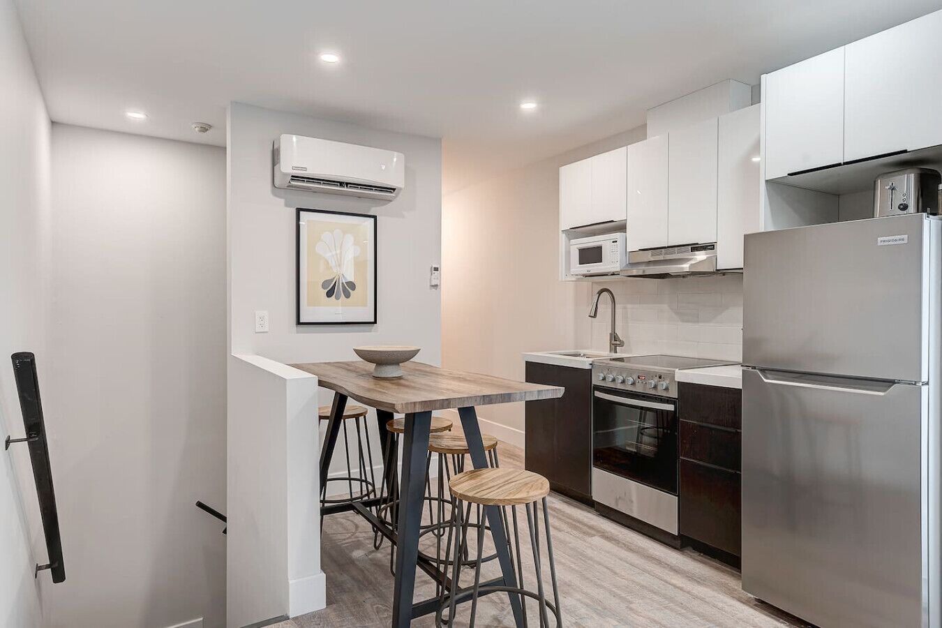 A compact kitchen with a built-in oven, microwave, and stainless steel refrigerator. There is a wooden table with stools and a framed picture on the wall.