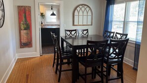 A dining area with a wooden table and chairs, a decorative wine bottle painting, a window with curtains, and a kitchen visible through an open door.
