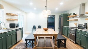 A modern kitchen with green cabinets, a wooden dining table, and a hanging light fixture.