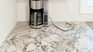 A kitchen with two wooden shelves holding white mugs, a coffee maker, and a grinder on a granite countertop.