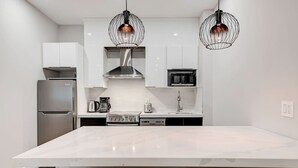 Une cuisine moderne avec des meubles blancs, un réfrigérateur en acier inoxydable, un four à micro-ondes, une machine à café et deux lampes suspendues.