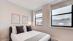 Une chambre à coucher avec un lit, deux œuvres d’art encadrées, une fenêtre donnant sur des immeubles et une lampe sur pied.