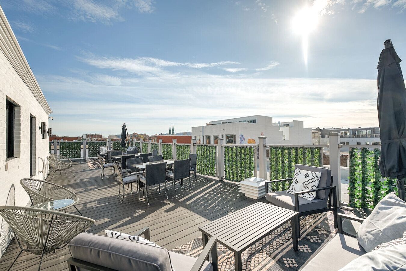 A rooftop terrace with outdoor seating, including chairs and tables, surrounded by a white building and greenery.