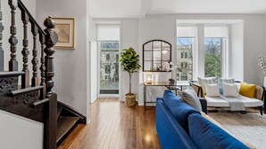Amplia sala de estar con un sofá azul, comedor con mesa y sillas, escalera con barandillas decorativas y ventanas grandes con vistas a la ciudad.
