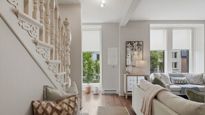Ein Wohnzimmer mit Treppe, einer Sofagarnitur, einem Beistelltisch mit Lampe und Blick durch ein Fenster.