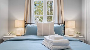 A bedroom with a large bed, two bedside lamps, a window with curtains, and a stack of towels on the bed.