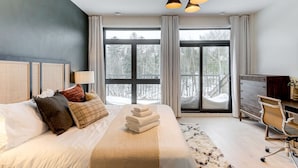 Ein modernes Schlafzimmer mit einem großen Bett, einem Schreibtisch und Blick auf einen verschneiten Wald durch große Fenster.