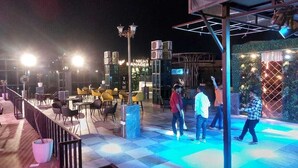 An outdoor seating area with tables and chairs, a bar area with a 'Night Manager' sign, and a decorative wall with lights. - Syrus Hotel  (Mathura)