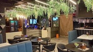 A modern indoor seating area with tables and booths, a bar area with screens, and hanging plants. - Syrus Hotel  (Mathura)