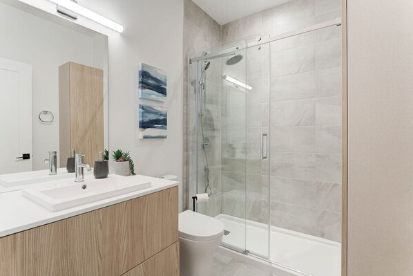 Un baño moderno con ducha envuelta en vidrio, un lavabo blanco con dos grifos y un mueble de tocador de madera.
