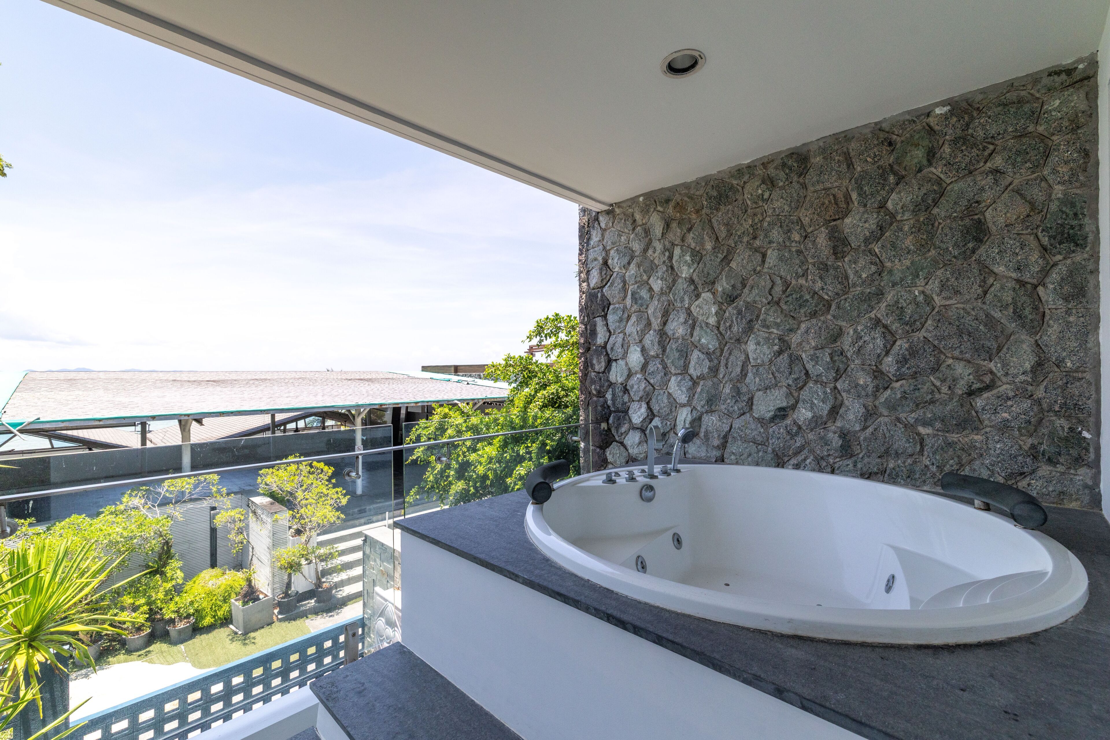 A modern bathroom with a stone wall, a large bathtub, and a balcony overlooking a garden and buildings.