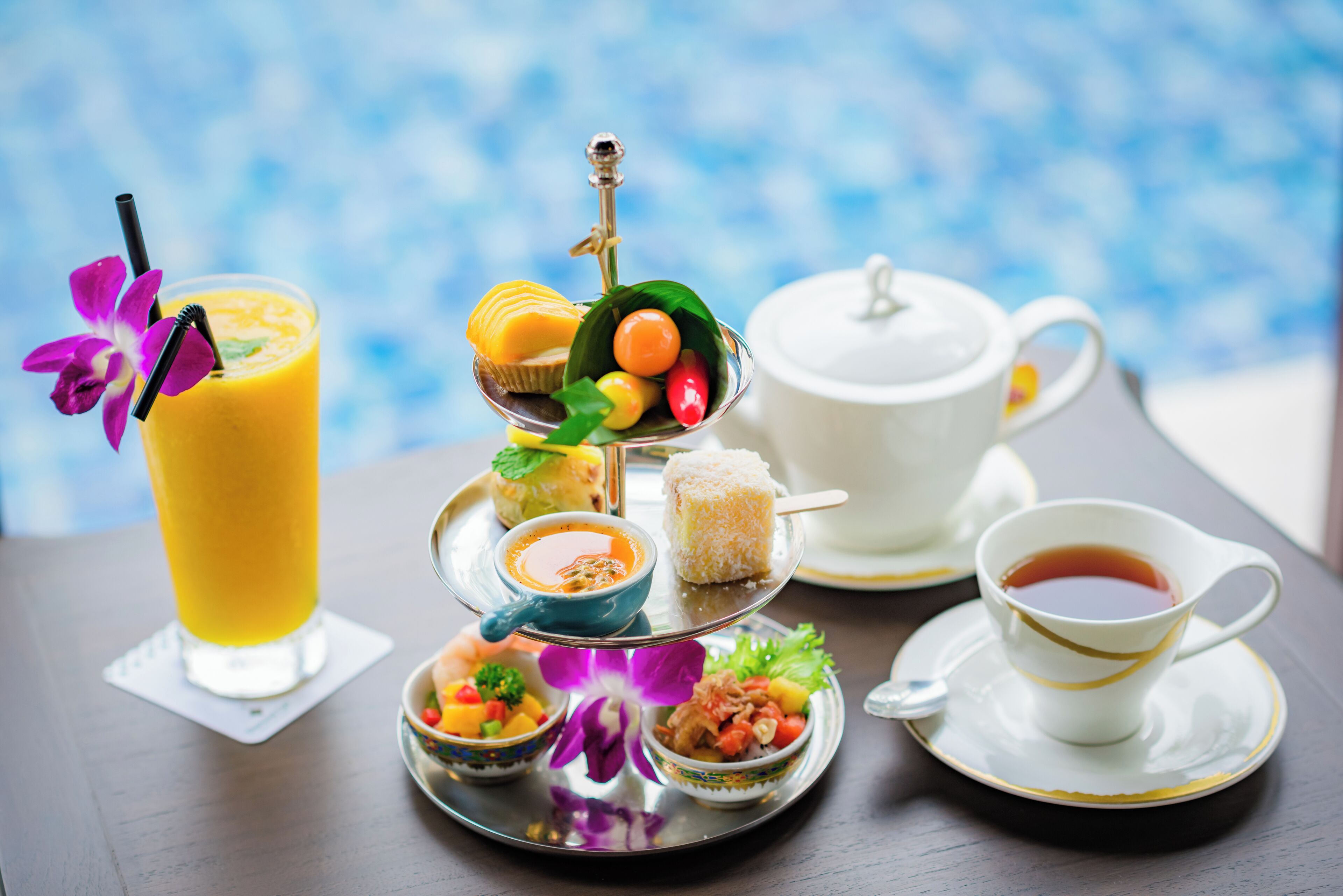 Bandeja escalonada com diversos alimentos e uma chávena de chá, colocada sobre uma mesa com uma chaleira branca e um copo de sumo de laranja.