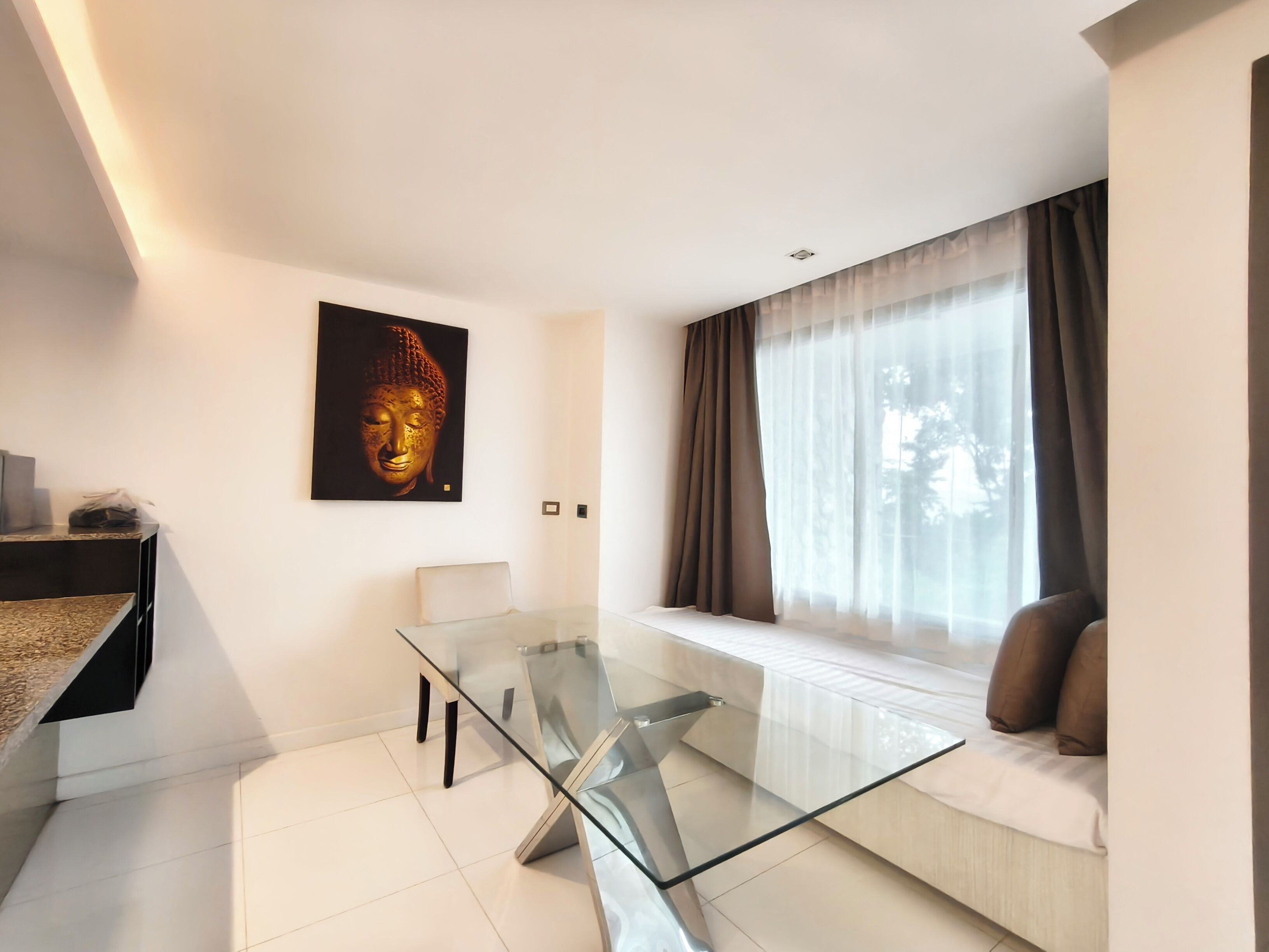 A modern living room with a glass dining table, a sofa with cushions, a Buddha statue, and a large window with curtains.
