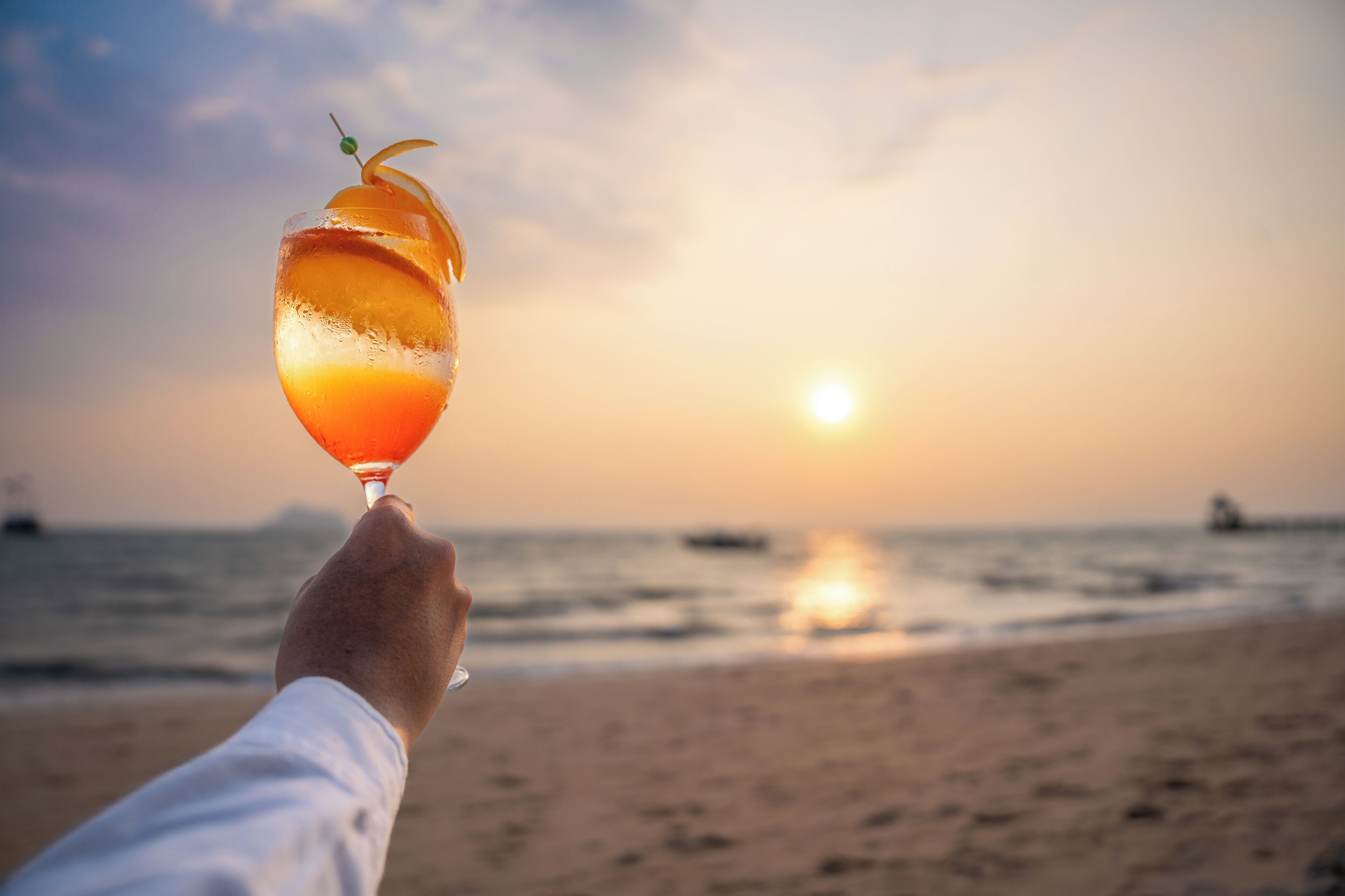 A hand holding a cocktail glass with a citrus garnish against a sunset beach backdrop.
