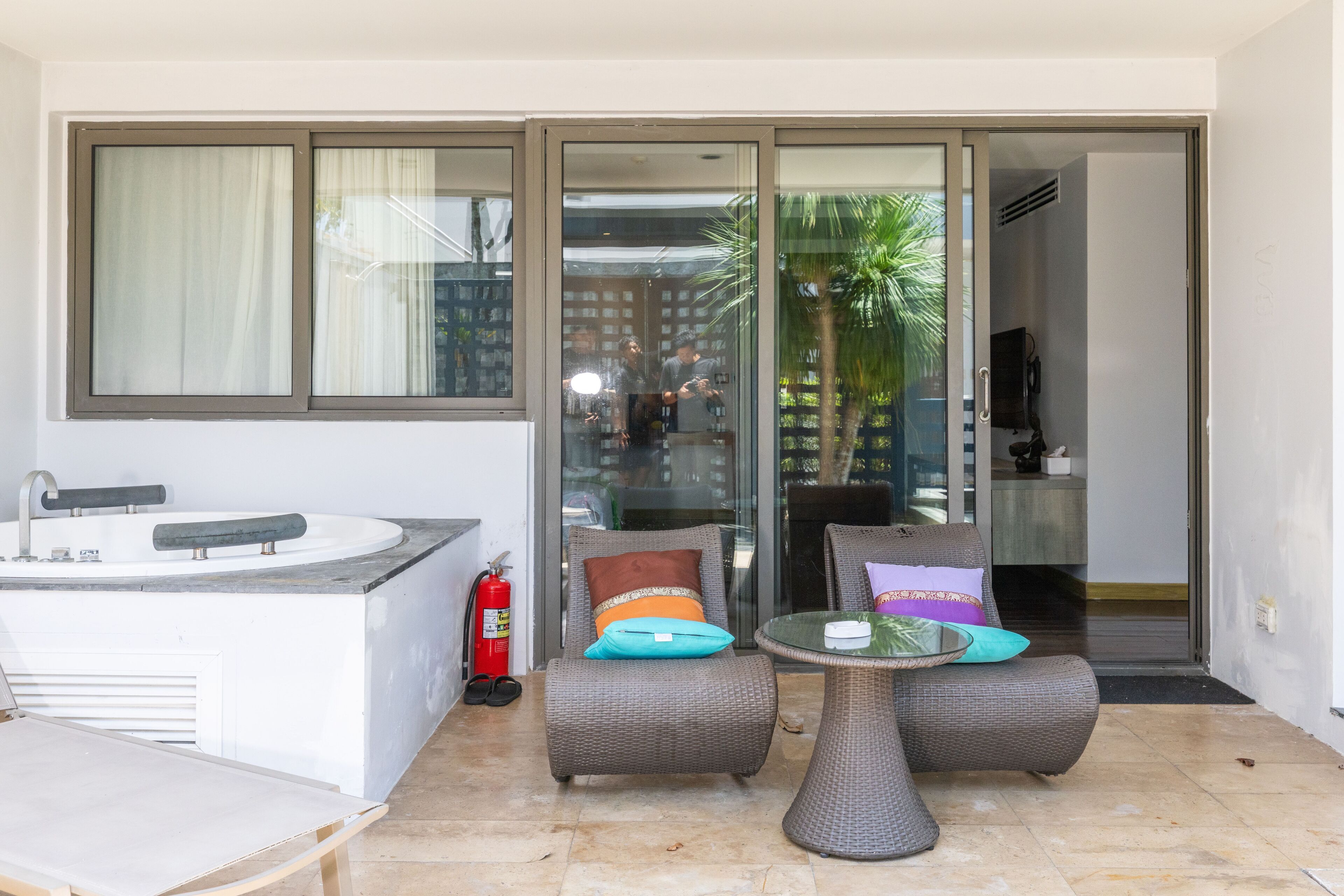 A modern bathroom with a sink and a toilet, a glass door leading to an outdoor area with two wicker chairs and a small table, and a fire extinguisher near the door.