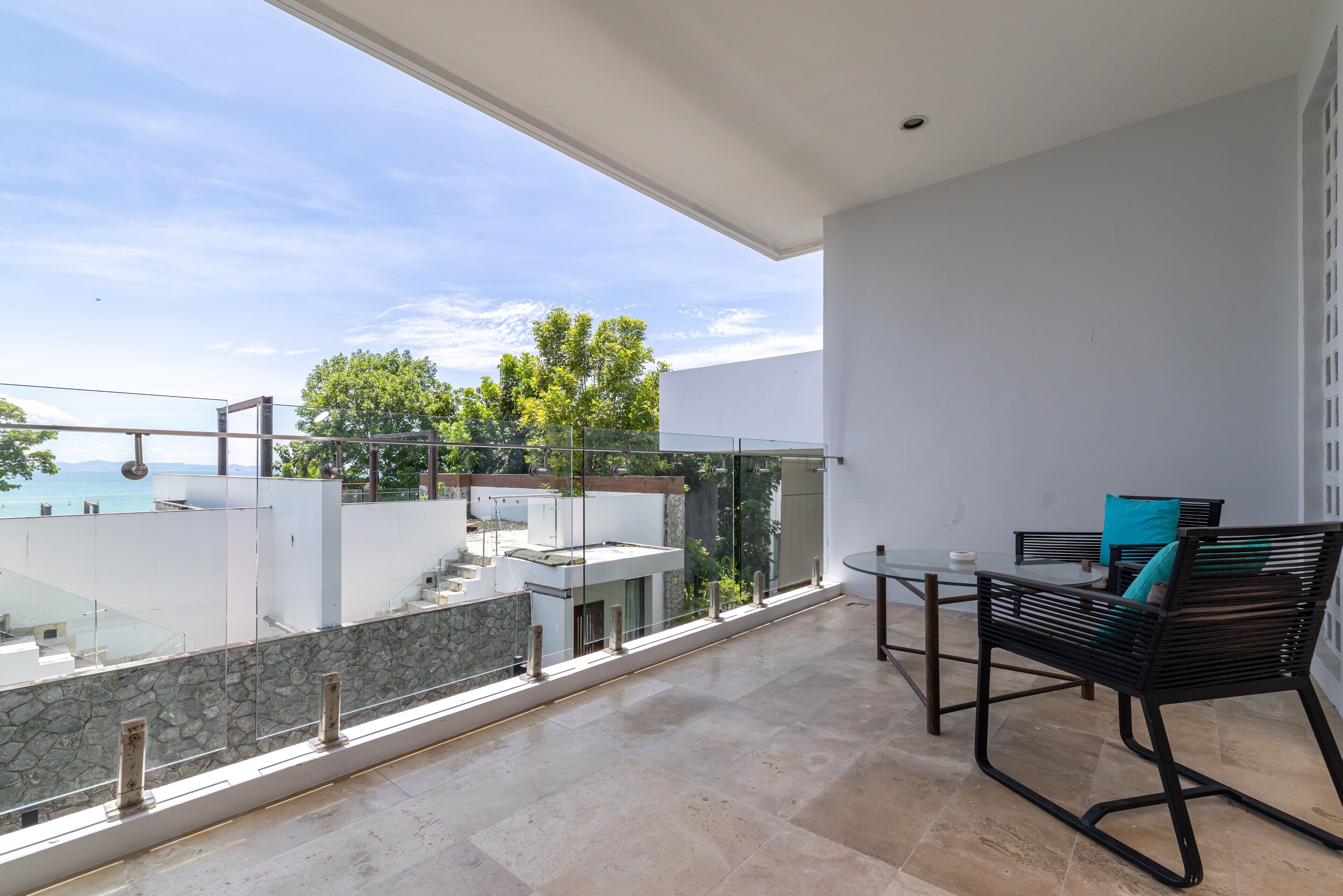 Un balcone con ringhiera in vetro, un tavolo con sedie e vista sul mare e sugli alberi.