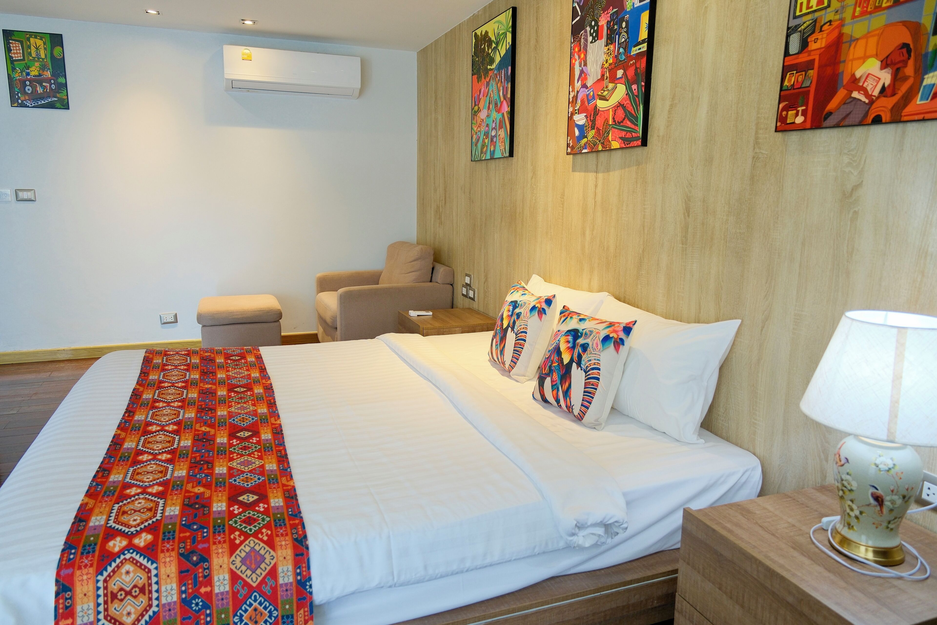A hotel room with a large bed, a bedside table with a lamp, a chair, and colorful artwork on the walls.