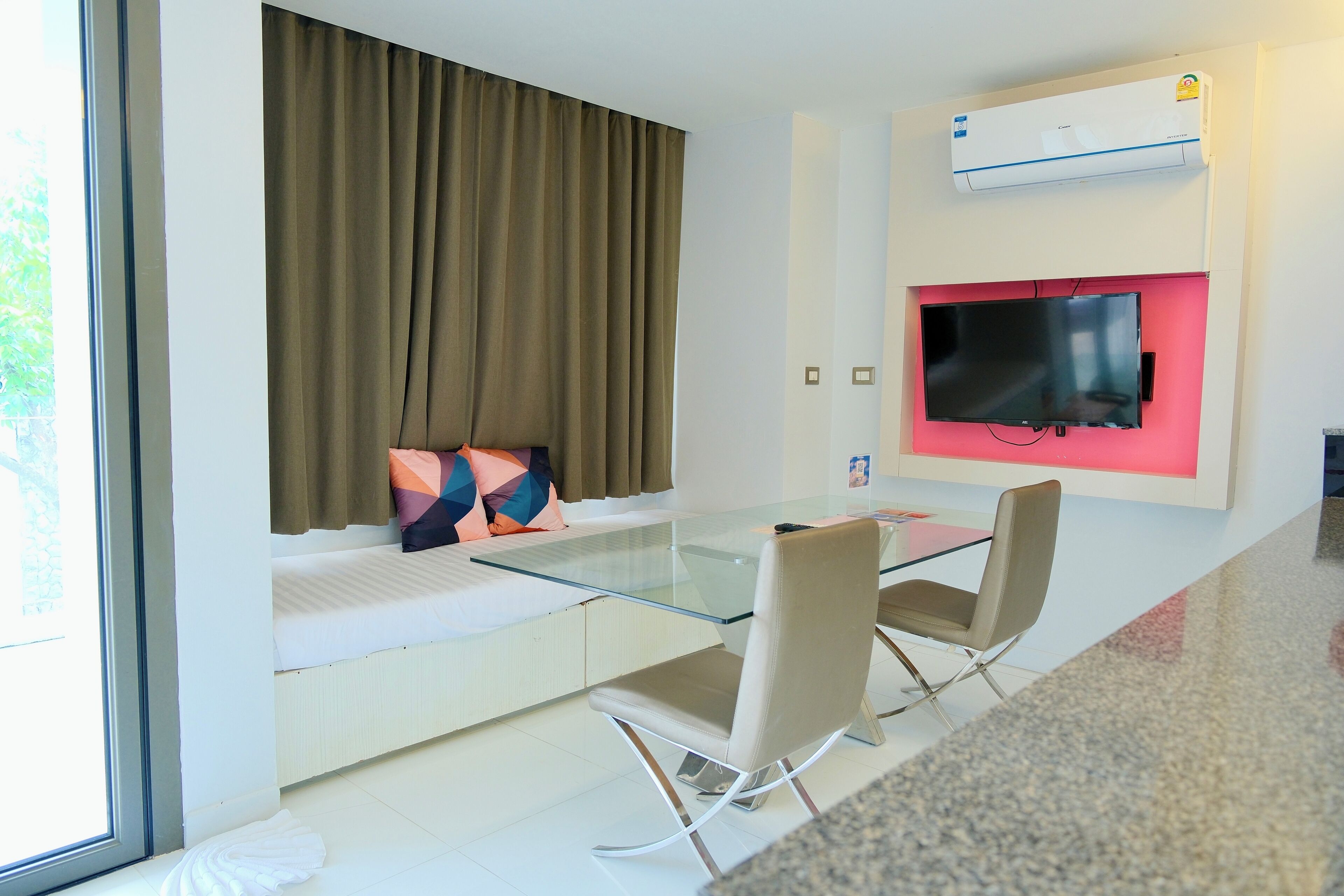 Una habitación de hotel moderna con una mesa de comedor de vidrio, dos sillas, un sofá y un televisor montado en la pared, además de un equipo de aire acondicionado.