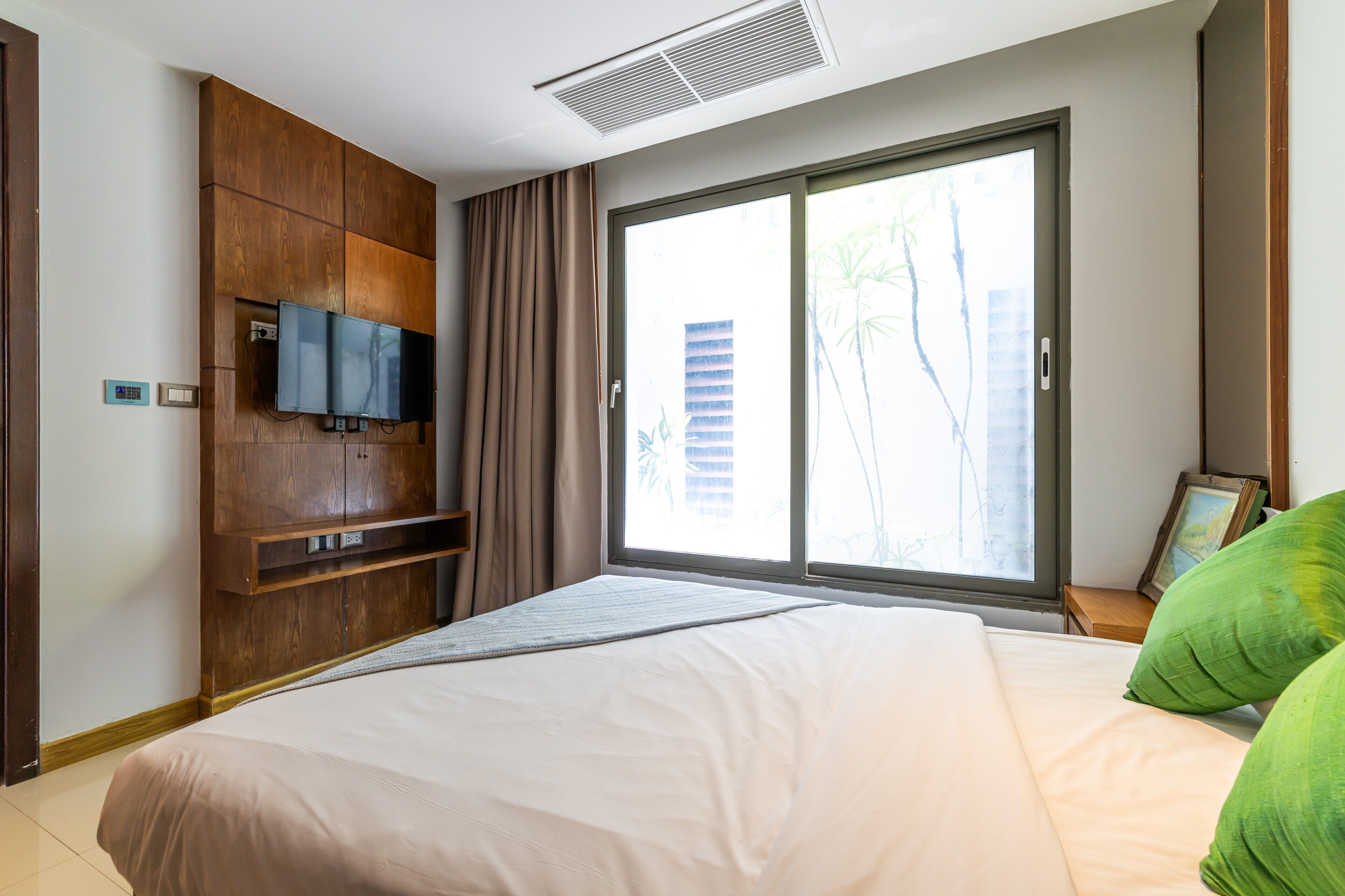 A hotel room with a large bed, a TV mounted on a wooden cabinet, a window with a view of buildings, and a small table with a picture frame.