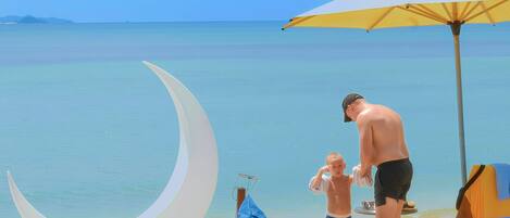 Escena de playa con una estructura en forma de luna creciente, una sombrilla amarilla y un niño y un adulto cerca del agua.