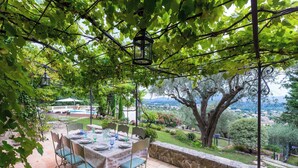 Ein Essbereich im Freien mit Tisch für vier Personen, umgeben von Stühlen, unter einer Pergola mit hängenden Laternen. Eine Steinmauer begrenzt den Bereich, und es ist eine Sicht auf eine entfernte Landschaft zu sehen.