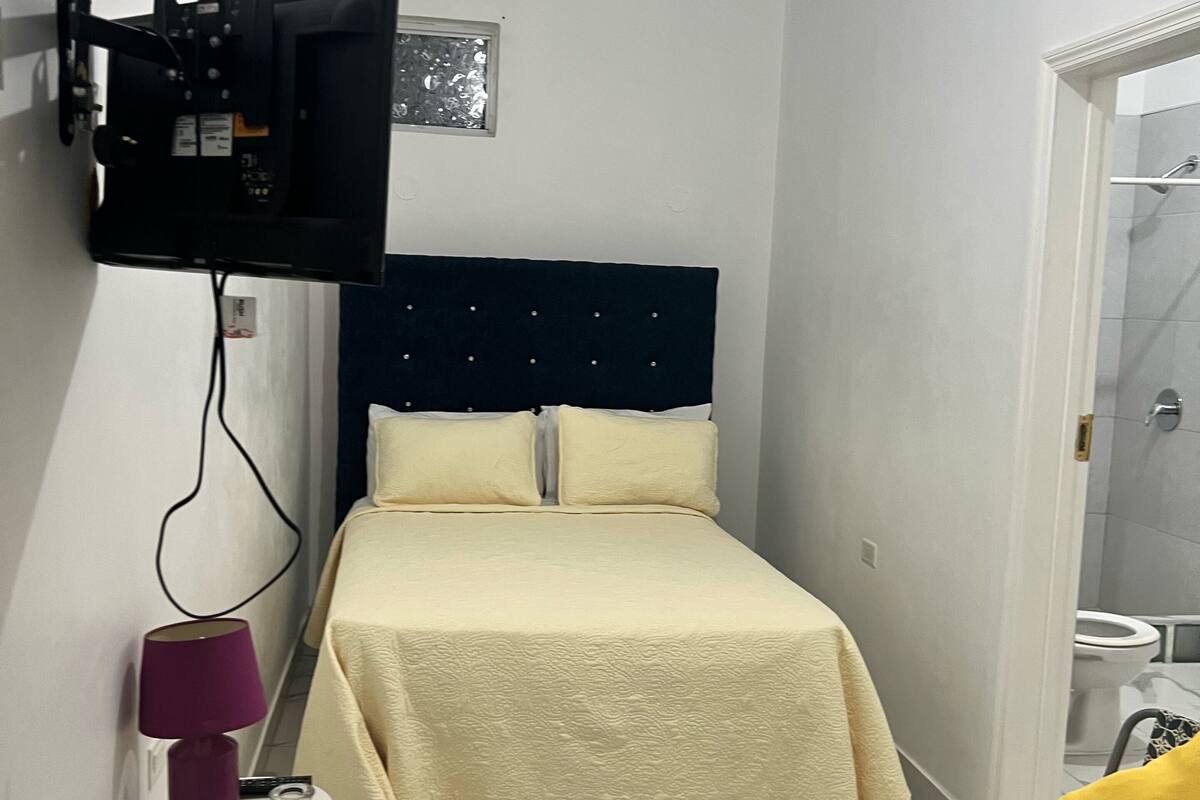 Un dormitorio con una cama, un televisor montado en la pared, una silla y un baño visible a través de una puerta abierta.