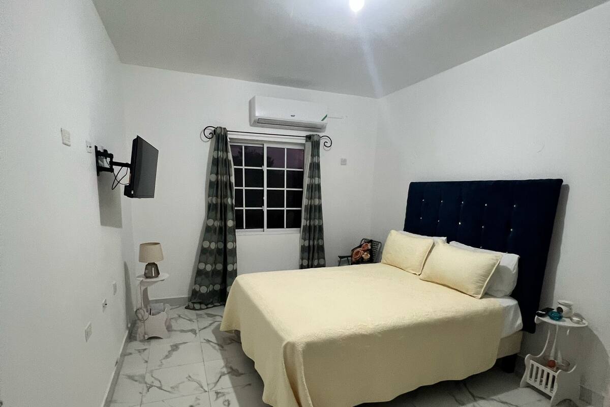 Un dormitorio con cama, televisor montado en la pared, ventana con cortinas y aire acondicionado.