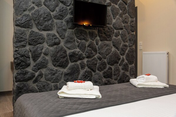 Une chambre d’hôtel avec un mur en pierre, un téléviseur à écran plat fixé au-dessus du lit, et deux serviettes soigneusement pliées posées sur le lit.