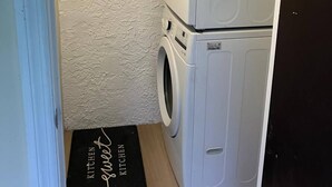Ein Waschraum mit gestapelten Waschmaschine und Trockner, einer strukturierten Wand und einer schwarzen Matte mit der Aufschrift 'Kitchen Sweet Kitchen'.